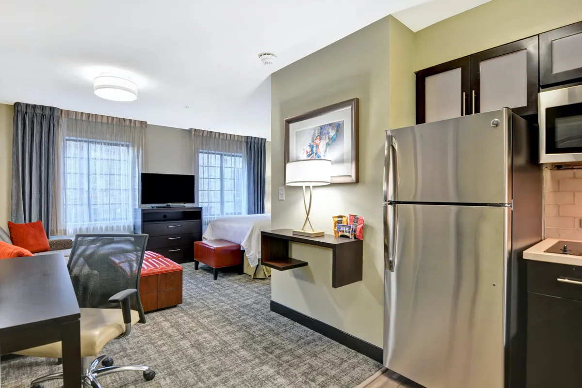 The hero image showcases a thoughtfully designed studio suite featuring a cozy bed, a modern kitchenette ideal for preparing simple meals, and a comfortable seating area perfect for relaxing or watching TV. Natural light bathes the space, creating an inviting and homey atmosphere ideal for couples or solo travelers.
