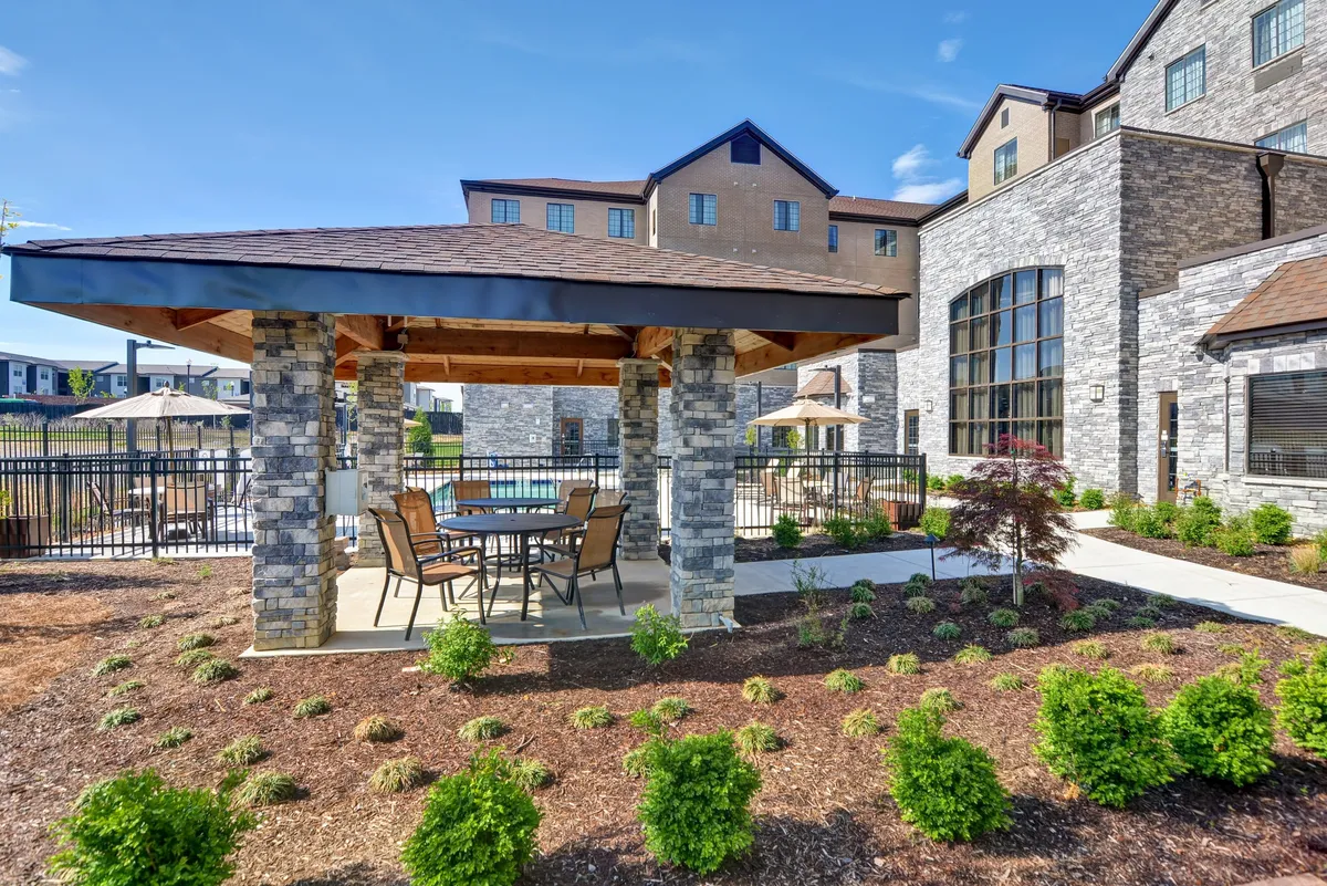This outdoor scene captures a stone gazebo with a table and chairs, set amid landscaped gardens and with a swimming pool visible in the background. The space is set up for outdoor dining, social gatherings, or quiet relaxation. The composition includes both greenery and recreational features, highlighting accessible leisure activities.