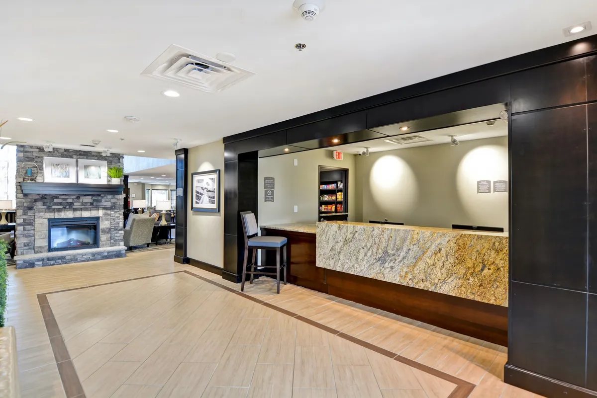 The image captures a welcoming front desk area with a cozy fireplace and a comfortable seating area. Guests are seen checking in, while others relax or socialize by the fire, emphasizing a friendly, inviting atmosphere reminiscent of both home and hotel hospitality.