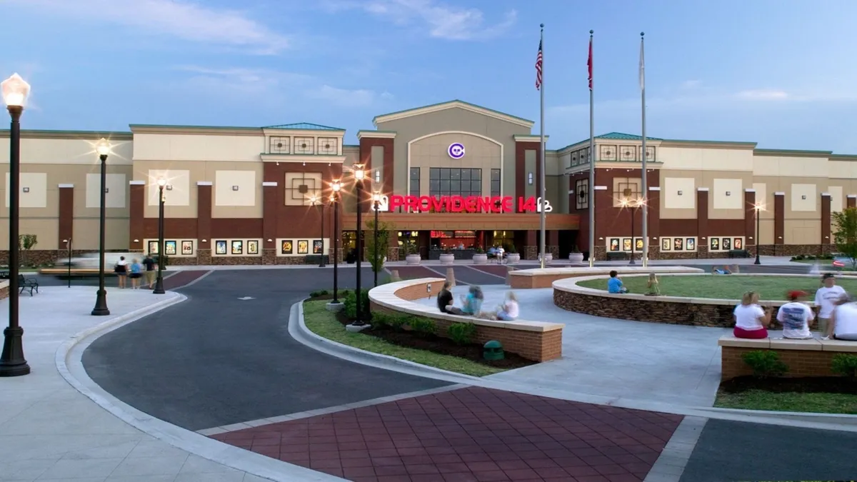 This image features the main entrance of the building, illuminated signage, and a landscaped seating area. There is an inviting outdoor environment where people can be seen socializing, possibly before or after catching a movie or enjoying a night out. The atmosphere is vibrant and welcoming, suggesting easy walkability to entertainment and dining options.