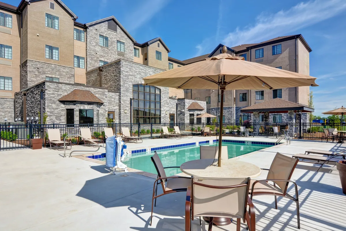 An inviting outdoor pool area with blue water, lounge chairs, umbrellas, and a visible hotel facade. The mood is relaxed and cheerful, suggesting a space ideal for swimming, sunbathing, and poolside gatherings.