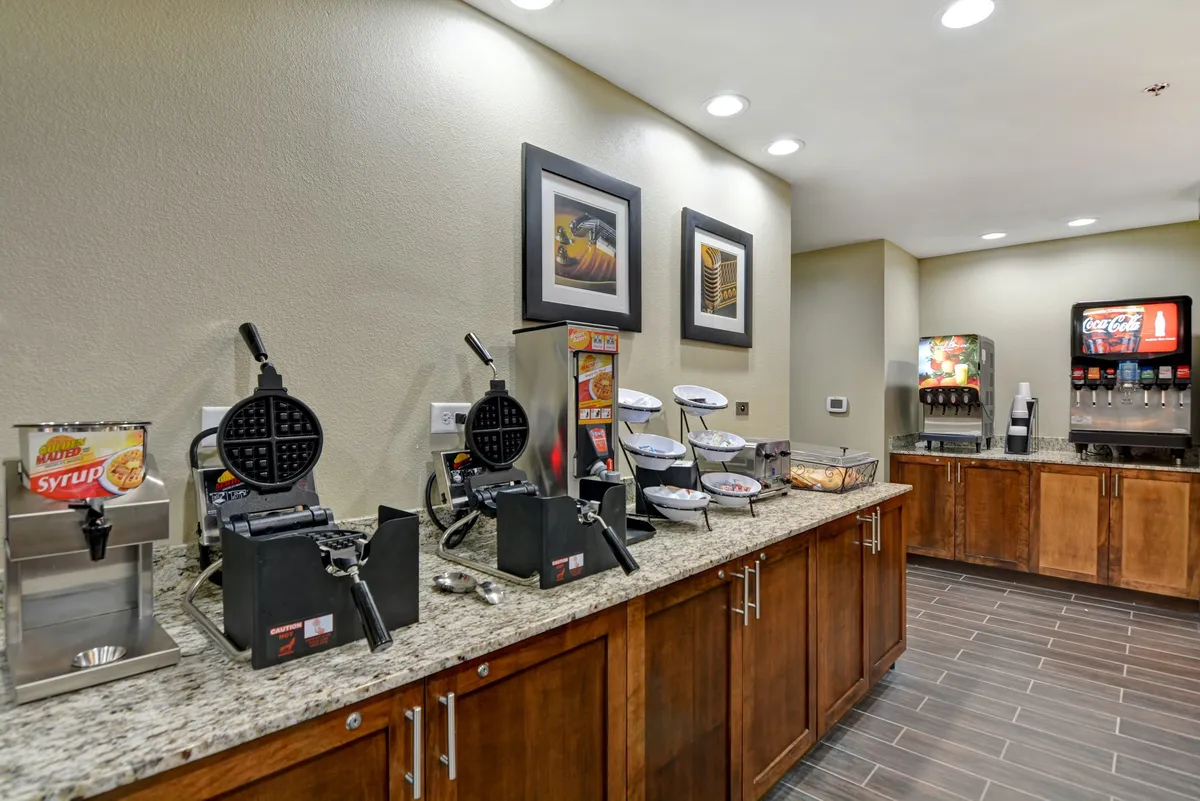 The image displays a breakfast area featuring waffle makers, a syrup dispenser, a beverage station, and food display trays. It captures a functional cooking and prep space, evoking the flexibility of preparing meals or snacks at one's convenience.