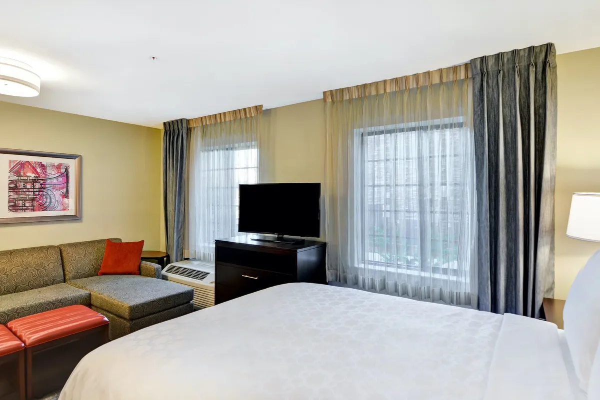 This image shows a well-appointed living space centered around a plush bed with crisp linens, flanked by a comfortable sofa and a flatscreen TV. Large windows allow natural light to fill the room, and tasteful artwork adds a unique touch to the décor. The workspace is subtly integrated, making the area suitable for both relaxation and productivity.
