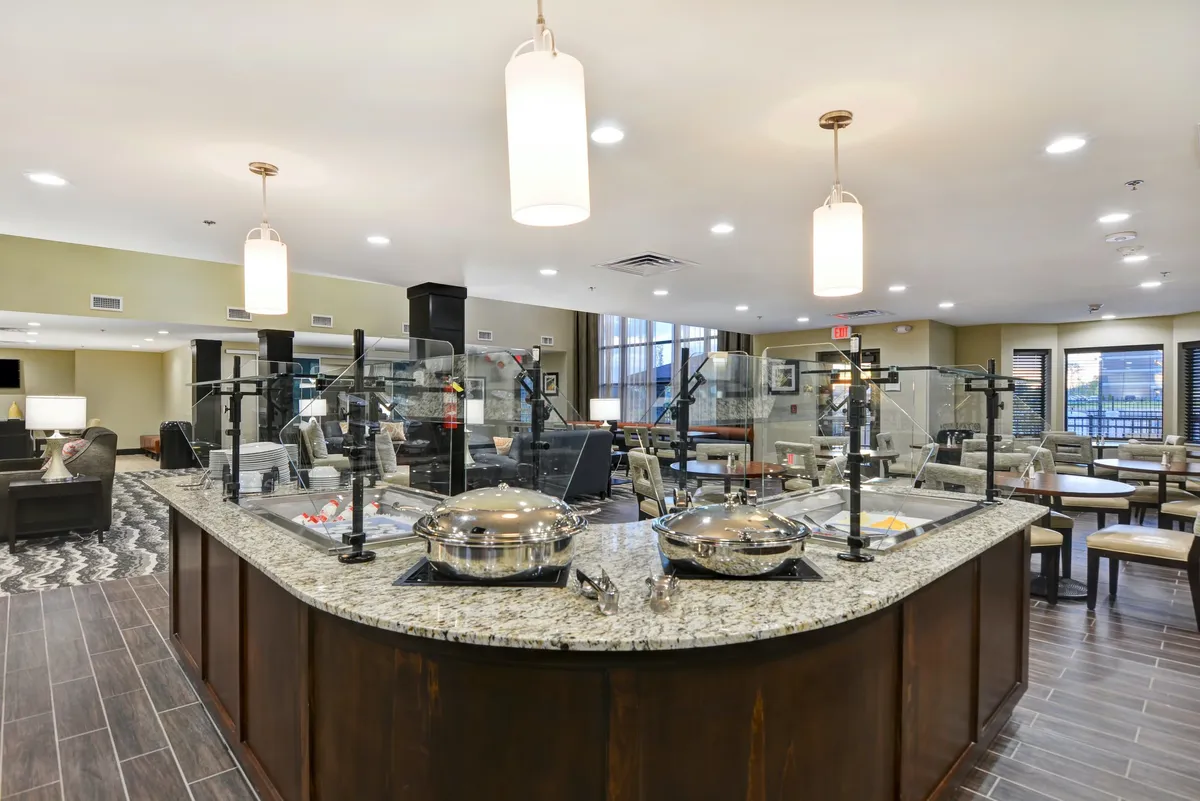 This image features a modern kitchenette area with a granite countertop, serving stations, and a comfortable seating area illuminated by natural light streaming through large windows. The space is designed to accommodate meal preparation and casual dining, with ample surfaces and storage visible.