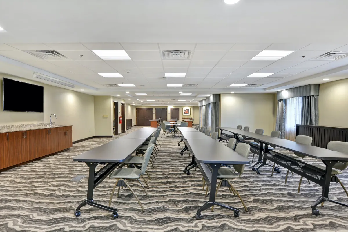 A business center featuring long tables set up for meetings, a wall-mounted TV, and a refreshment counter. The space has a focused yet collaborative feel, ideal for work or small professional gatherings.