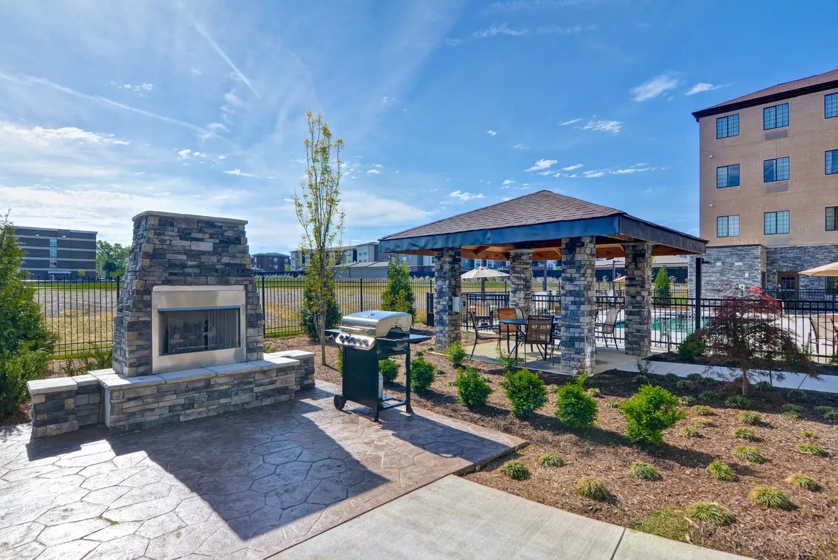This image shows an exterior gathering space featuring a stone fireplace, grill, covered gazebo, and lush landscaping. The area is designed for outdoor cooking, communal meals, and cozy evenings, indicating a lively community atmosphere and opportunities for spontaneous activities.