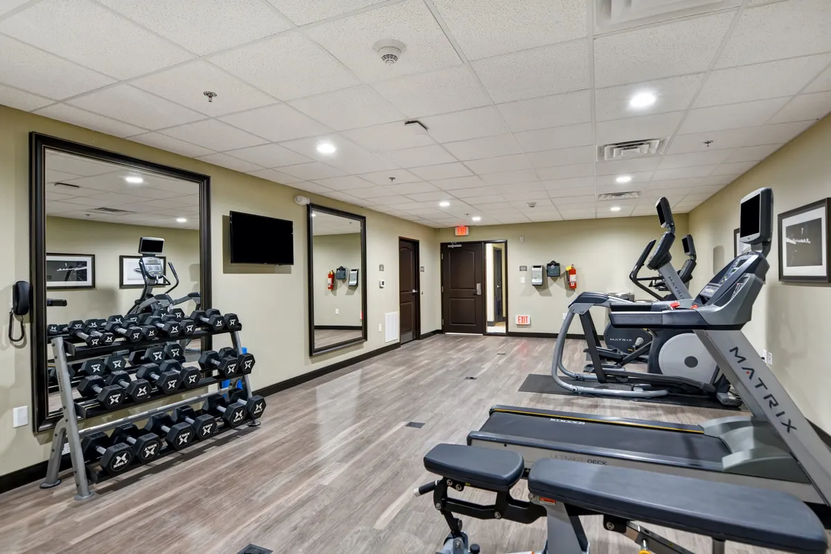The fitness center is equipped with a dumbbell rack, treadmills, ellipticals, and mirrors. The environment conveys an energetic mood, with several people engaged in workouts and group fitness activities.