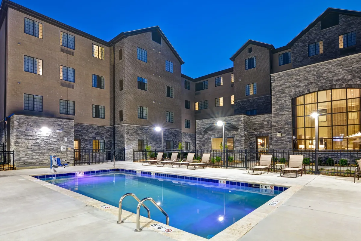 This image showcases the inviting exterior of Staybridge Suites, with a sparkling swimming pool, comfortable lounge chairs, and the building facade visible in the background. Outdoor lighting enhances the welcoming atmosphere, making the space feel open and connected to the outside world.