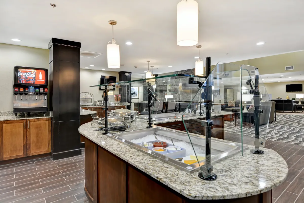 This image displays a granite countertop arranged with a well-stocked breakfast buffet, complemented by a Coca-Cola dispenser and illuminated by hanging lights. The setup is inviting and conducive to social self-service dining, with a casual and welcoming atmosphere.